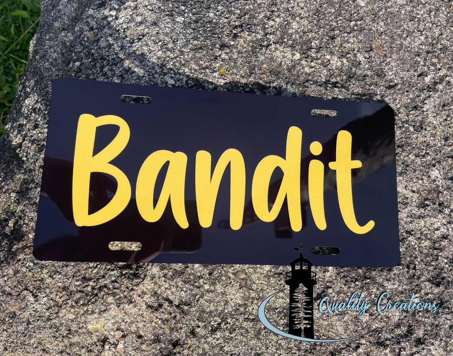 A black novelty license plate with the word 'Bandit' in large yellow script, photographed resting on a textured gray stone surface outdoors.