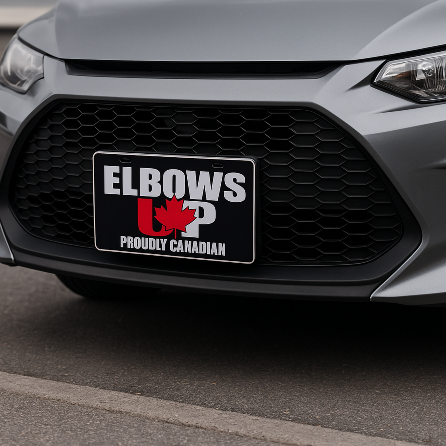 Front view of a silver car with an “Elbows Up Proudly Canadian” license plate mounted on the bumper, featuring a red maple leaf design, photographed outdoors during the day.