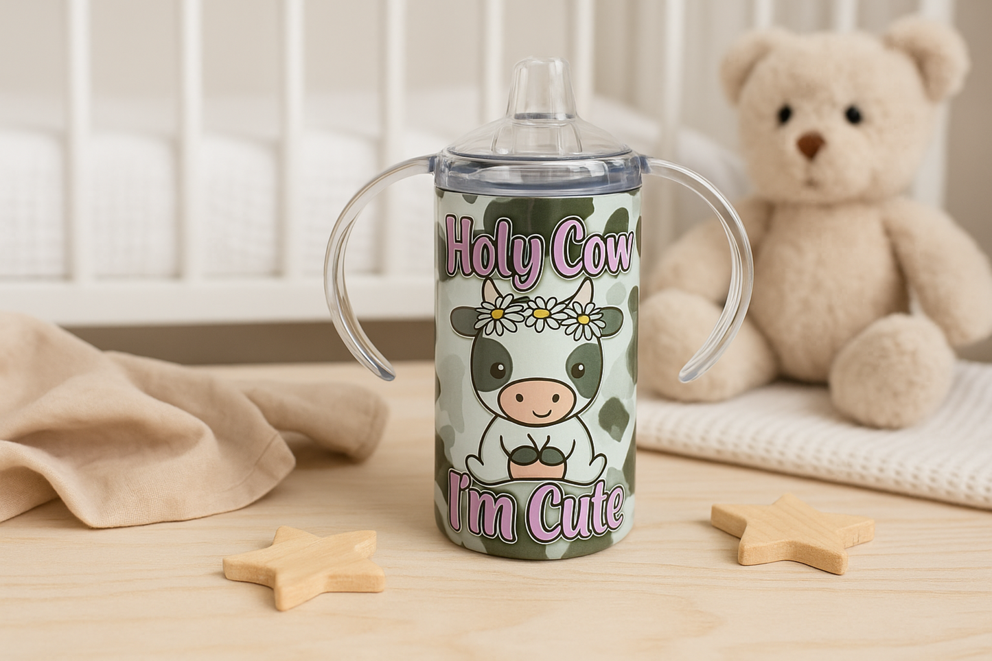 Cow-themed sippy cup with handles shown on a nursery table with a teddy bear, wooden toys, and a soft beige blanket