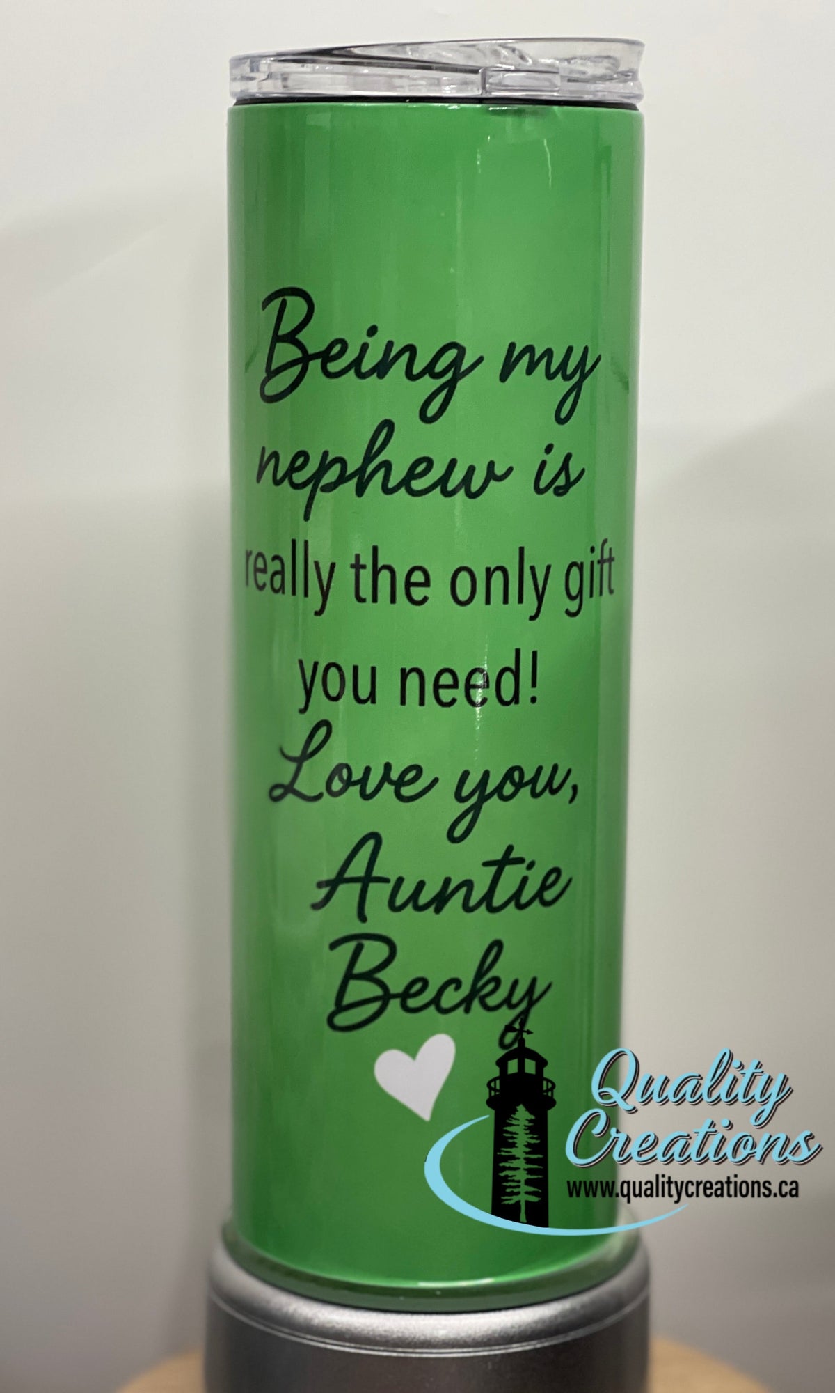 A green insulated tumbler with a clear lid, printed with the phrase 'Being my nephew is really the only gift you need! Love you, Auntie Becky' in black script. A small lighthouse and heart graphic appears near the bottom, along with a 'Quality Creations' watermark/logo.