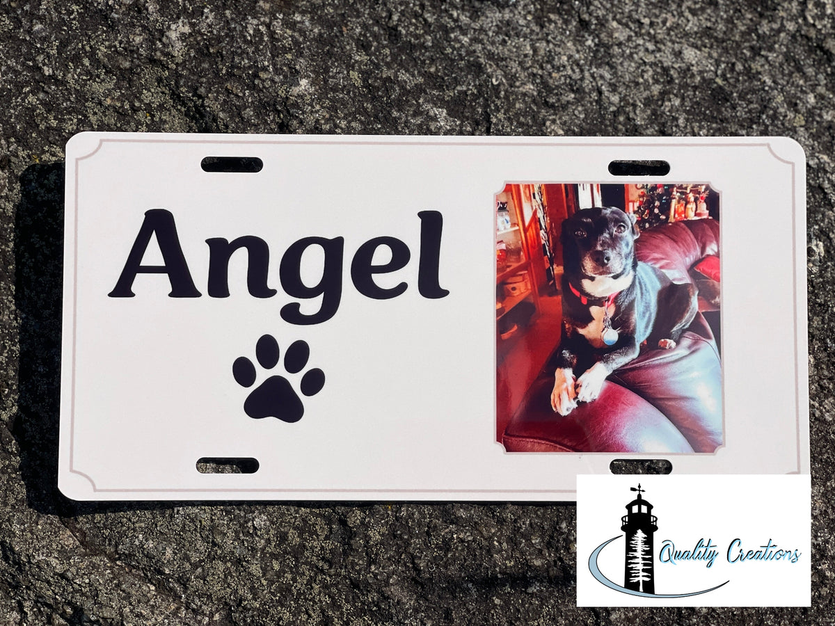 A white custom license plate featuring the name 'Angel' in black script, a black paw print, and a rectangular inset photo of a dog on a red couch. The plate is photographed on a dark asphalt surface, with a 'Quality Creations' logo in the bottom right corner.