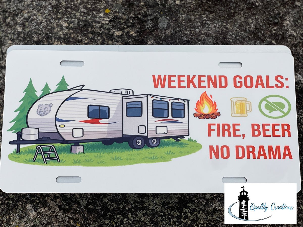 A novelty license plate featuring a camping trailer illustration and the text 'WEEKEND GOALS: FIRE, BEER NO DRAMA' with small icons of a campfire, a beer mug, and a crossed-out beer glass. The plate is photographed on a dark textured asphalt surface, with a 'Quality Creations' logo in the bottom right corner.