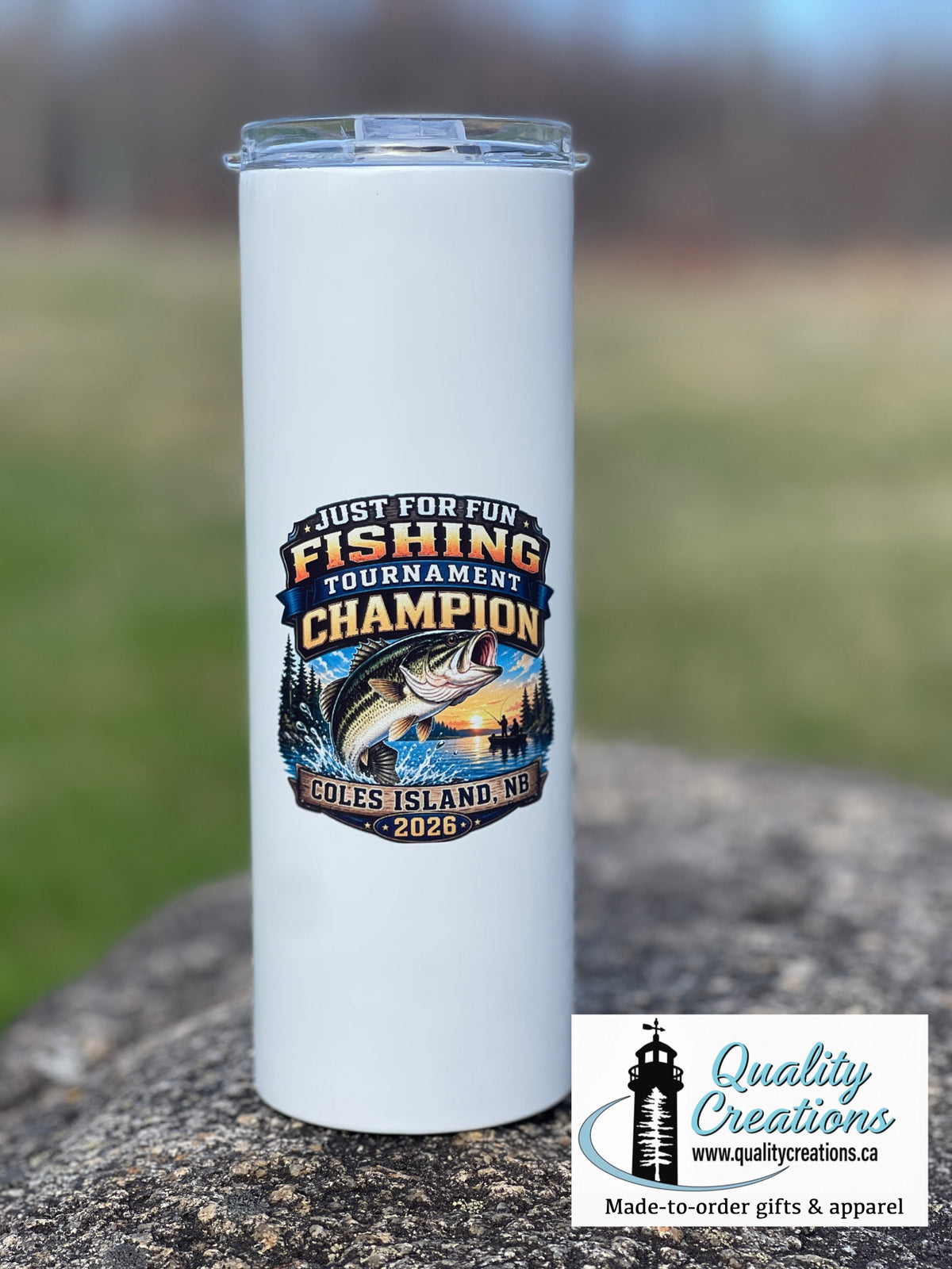 A tall white insulated 20oz tumbler with a clear screw in  lid sits on a stone surface outdoors. The tumbler has a large fishing-themed graphic reading "Just For Fun Fishing Tournament Champion" with a fish illustration and "Coles Island NB 2026" text.