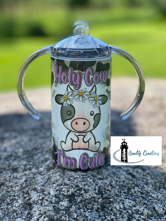 Cow-themed toddler sippy cup with “Holy Cow I’m Cute” text, dual handles, and floral cow graphic on a stone background