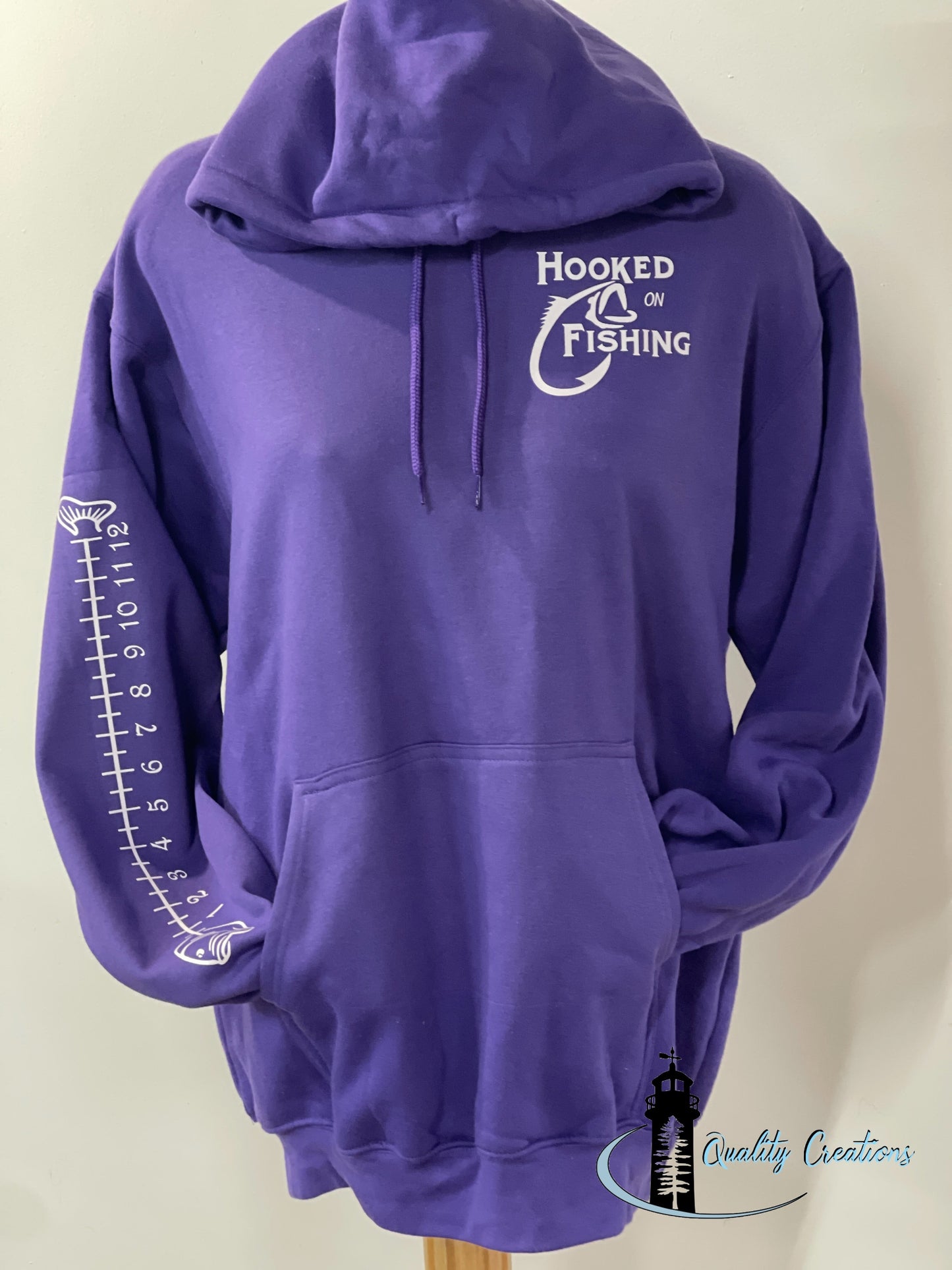 Purple hoodie with a white ‘Hooked on Fishing’ graphic on the chest and a vertical fish ruler design down the sleeve. Displayed on a mannequin against a light background with the Quality Creations logo in the corner. qualitycreations247, Moncton, Salisbury, Shediac, river, newbrunswick, nb, Ontario 