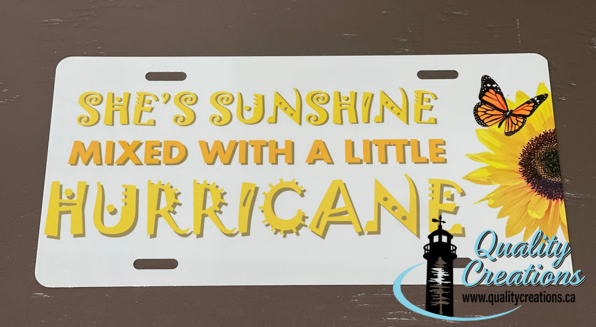 A novelty license plate with the text 'SHE'S SUNSHINE MIXED WITH A LITTLE HURRICANE' in yellow and orange lettering, featuring a yellow sunflower and an orange-and-black butterfly on the right side. The plate is photographed flat against a dark brown wooden surface, with a 'Quality Creations' watermark in the bottom right corner.