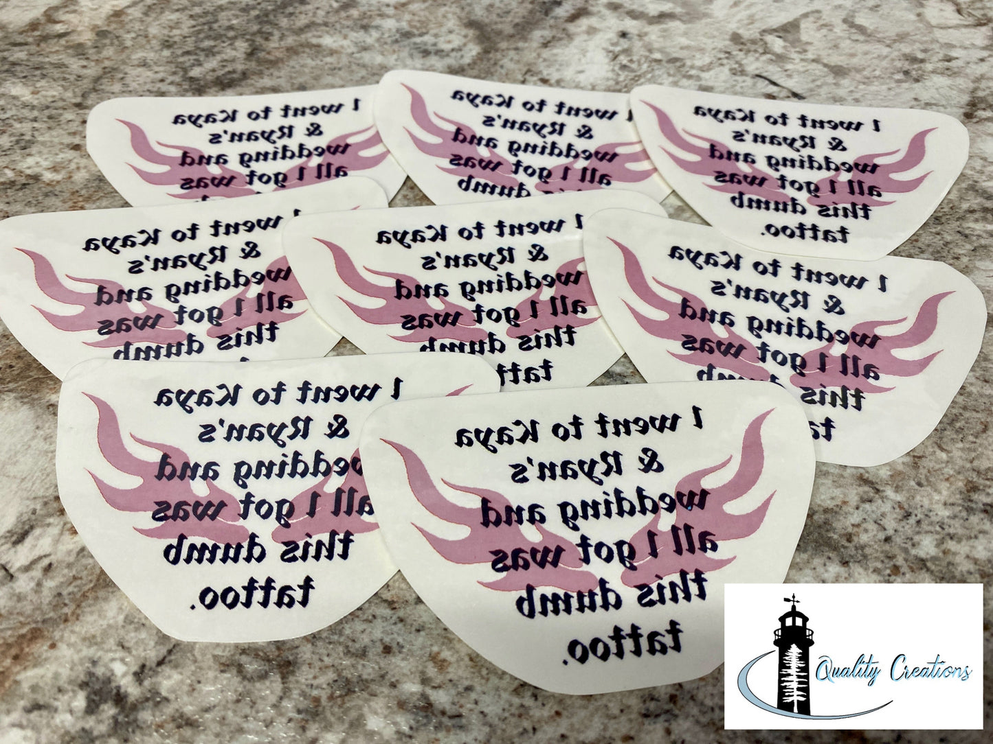 Six white tattoo sticker templates with pink wing designs and black text, arranged on a speckled granite countertop. A 'Quality Creations' logo is visible in the bottom right corner.