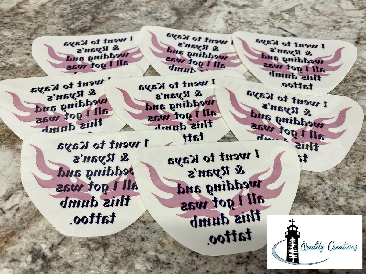 Six white tattoo sticker templates with pink wing designs and black text, arranged on a speckled granite countertop. A 'Quality Creations' logo is visible in the bottom right corner.