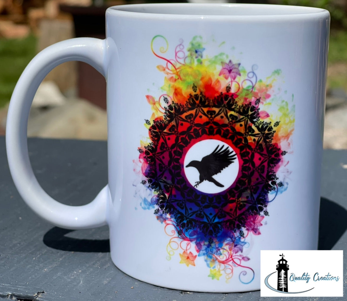 White mug featuring the Crow’s Nest logo with colourful mandala and black crow silhouette, photographed on a grey surface outdoors in natural light