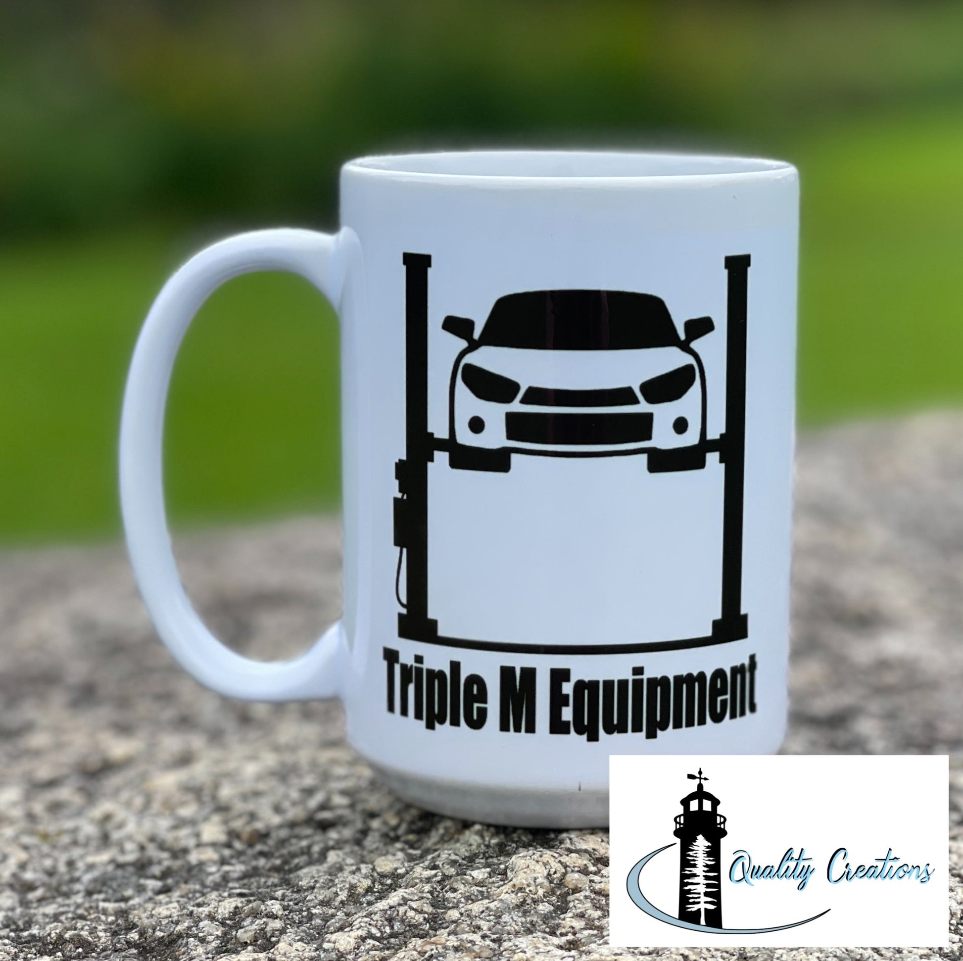 
White mug with Triple M Equipment logo featuring a lifted truck design, photographed outdoors on a rock with a green grass background