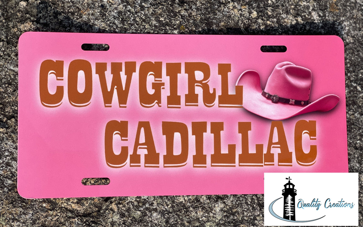 A pink novelty license plate featuring the words 'COWGIRL CADILLAC' in brown lettering and a pink cowboy hat illustration, photographed on a gray stone surface. A 'Quality Creations' logo appears in the bottom right corner.