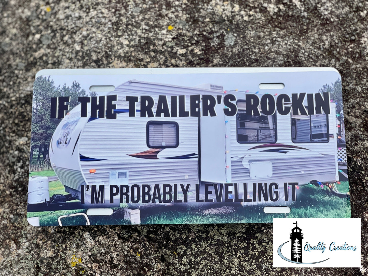 A novelty license plate featuring a printed photo of a travel trailer with bold text that reads 'IF THE TRAILER'S ROCKIN' and 'I'M PROBABLY LEVELLING IT', photographed on a textured gray stone surface. A 'Quality Creations' logo is visible in the bottom right corner.