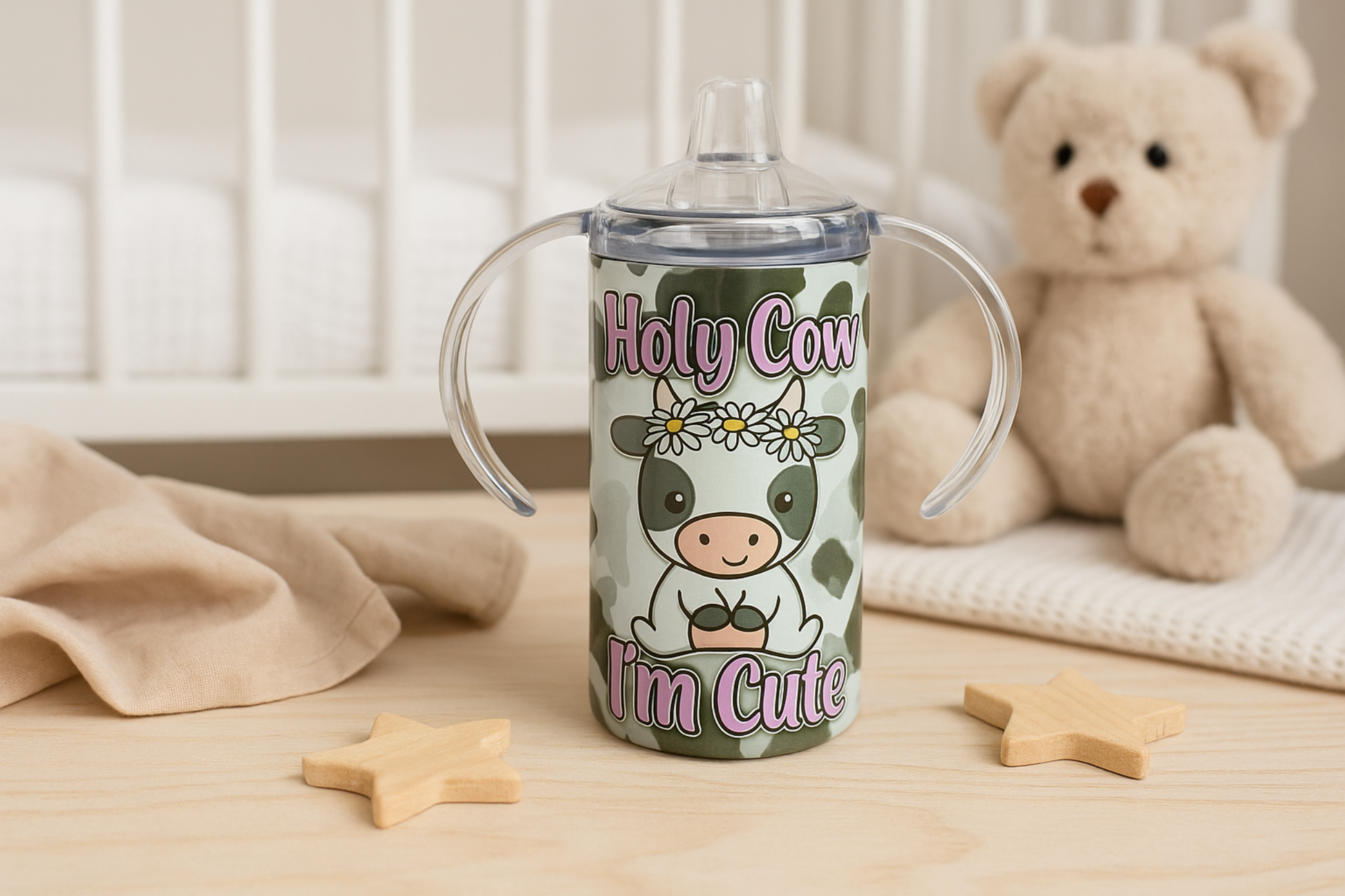 Cow-themed sippy cup with handles shown on a nursery table with a teddy bear, wooden toys, and a soft beige blanket