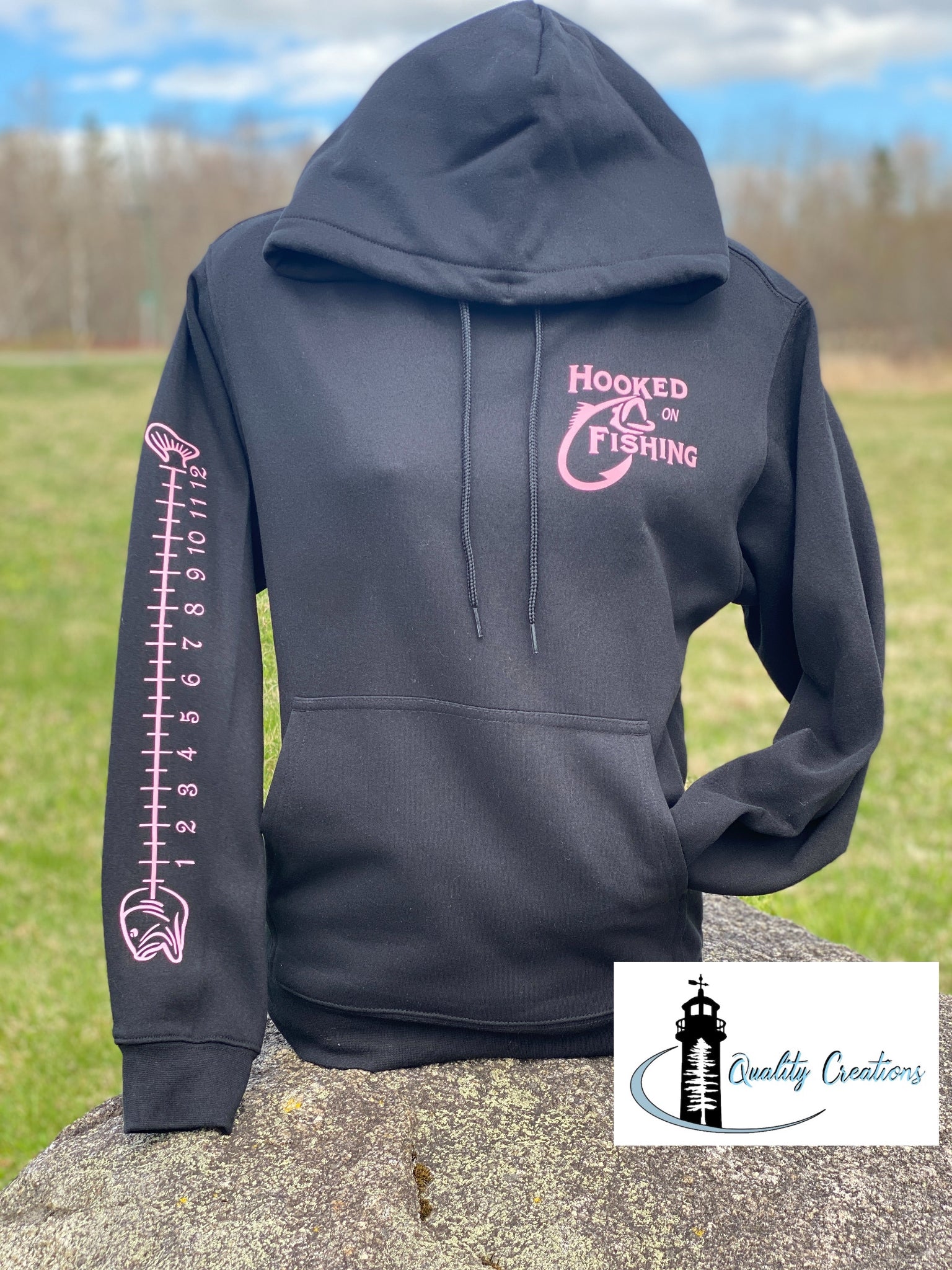 Black hoodie with a pink ‘Hooked on Fishing’ graphic on the chest and a vertical fish ruler design down the sleeve. Displayed on a mannequin against a light background with the Quality Creations logo in the corner. qualitycreations24, Salisbury, Canada, river, ocean, newbrunswick, nb, Ontario 