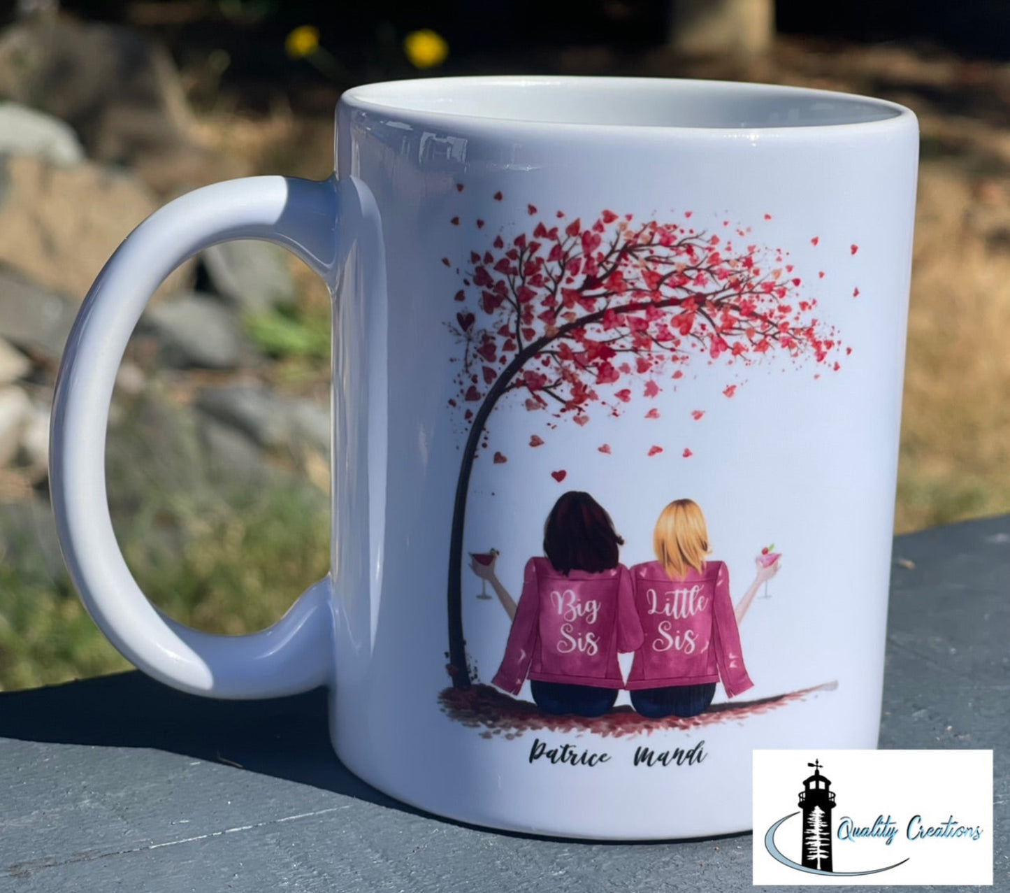 White mug with a custom design of two sisters in pink jackets under a red heart tree, shown outdoors on a rock