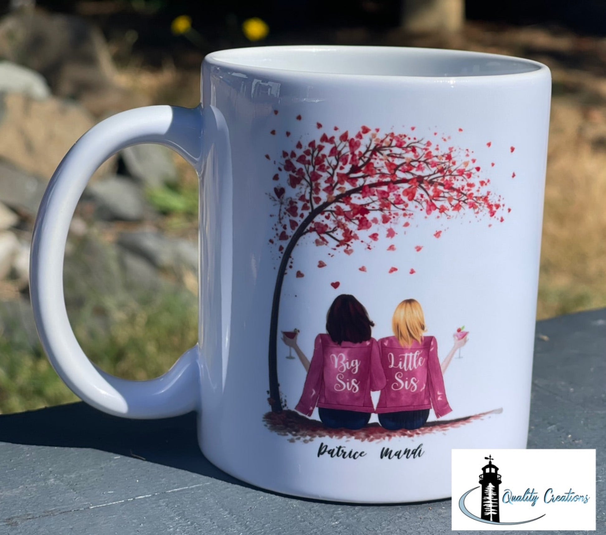 White mug with a custom design of two sisters in pink jackets under a red heart tree, shown outdoors on a rock