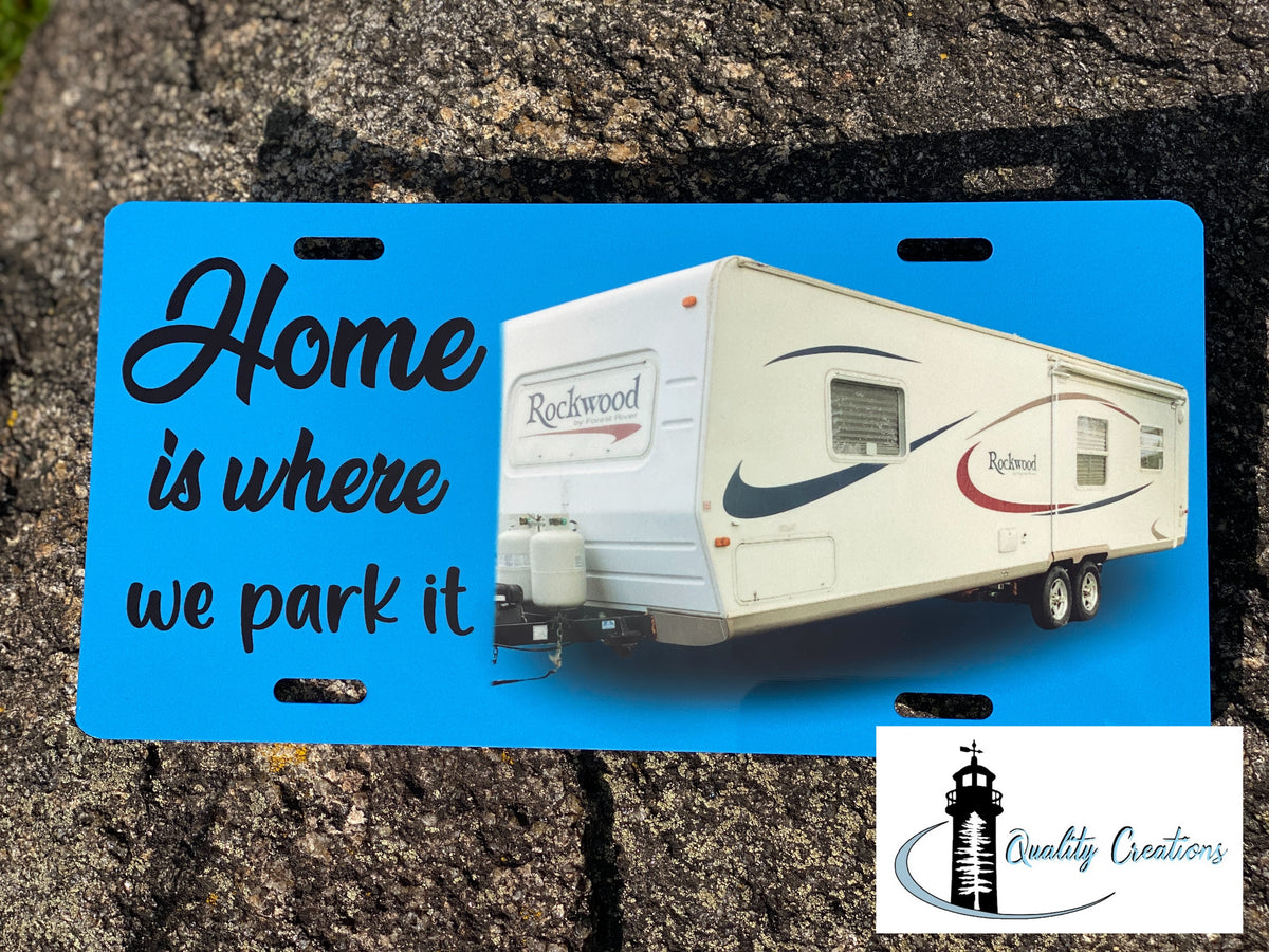 A novelty license plate featuring a bright blue background with the phrase 'Home is where we park it' in black script and serif text, alongside a printed photo of a white Rockwood travel trailer. The plate is photographed flat against a textured gray stone background, with a 'Quality Creations' logo watermark in the bottom right corner.