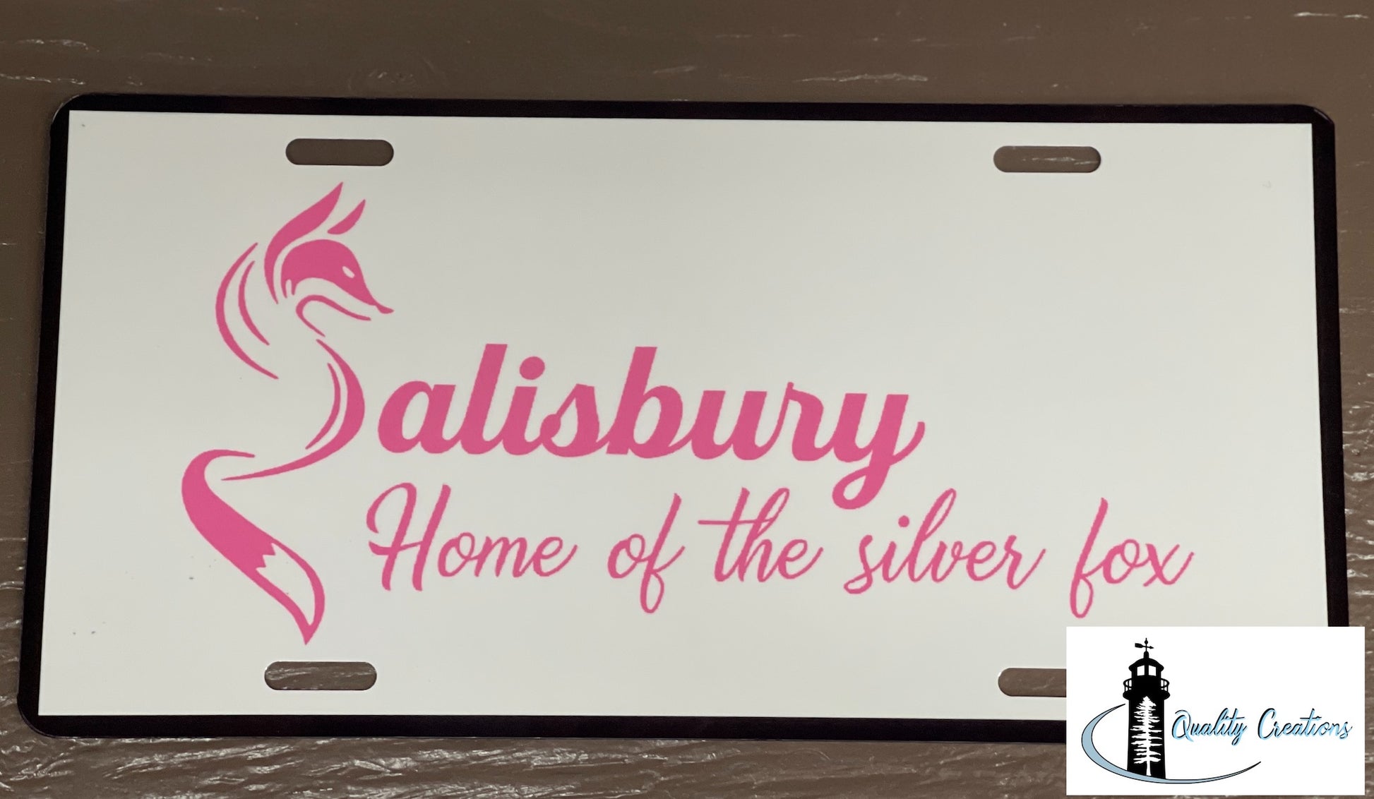 A white novelty license plate with a black border, featuring a pink fox graphic and the text 'Salisbury Home of the silver fox' in pink script. The plate is photographed flat on a dark brown wood-grain surface, with a 'Quality Creators' logo overlay in the bottom right corner.